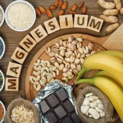 Magnesium-reiche Lebensmittel wie Sesam, Nüsse, Bananen und dunkle Schokolade sind auf einem Holztisch arrangiert.