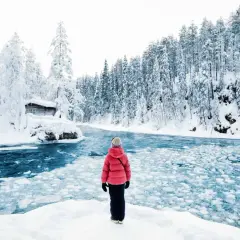 Une personne en manteau rouge admire un paysage hivernal de la pays Lapone, avec une rivière gelée et une cabane enneigée.
