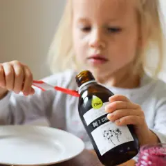 Mała dziewczynka trzyma butelkę z napisem "Olej z czarnuszki dla dzieci". Obok stoi talerzyk i słoiczek z czymś czerwonym.