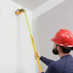 Mężczyzna w kasku mierzy ścianę za pomocą żółto-zielonego urządzenia na wysięgniku.