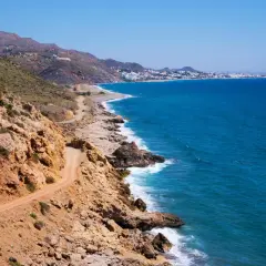 Costa rocosa con camino de tierra y el mar azul. Al fondo, se divisa la silueta de Mojácar y sus playas.