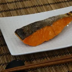A perfectly grilled salmon shioyaki rests on a white plate, next to a bowl of white rice and chopsticks.