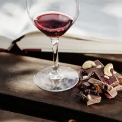 Un bicchiere di vino rosso accanto a pezzi di cioccolato con frutta secca e noci. Un perfetto abbinamento vino e cioccolato per una serata di relax.
