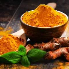 Radici di curcuma e polvere dorata in un cucchiaio e ciotola, con foglie verdi. La curcuma è nota per le sue proprietà antinfiammatorio.