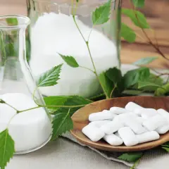 Ksylitol co to? Biały proszek w kolbie i słoiku, obok drewnianej łyżki z pastylkami. Liście brzozy sugerują naturalne pochodzenie.