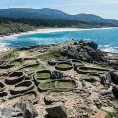 Ruinas circulares de piedra, vestigios de antiguos poblados, se extienden sobre un promontorio rocoso con el mar azul al fondo. Estos son algunos de los monumentos de Galicia.