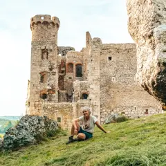 Kobieta siedzi na trawie przed ruinami zamku, idealne miejsce na postój podczas podróży szlakiem orlich gniazd samochodem.