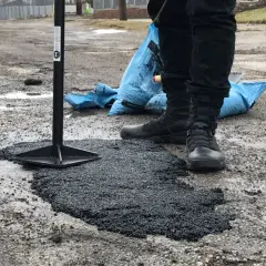 Pracownik ubija asfalt na zimno, naprawiając dziurę w drodze. Obok leży niebieski worek z materiałem.