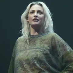 Blond wokalistka w siatkowej bluzce, przypominająca styl wokalisty Linkin Park, na ciemnym tle.