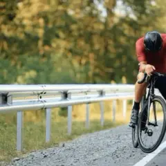 Cycliste en tenue de sport, casque noir, s'appuyant sur son vélo sur le bord de la route. Il semble fatigué, peut-être après avoir fait du sport à jeun.