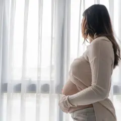 Kobieta w ciąży stoi przy oknie, patrząc na zewnątrz. Jej dłonie delikatnie obejmują brzuch, a w jej postawie można wyczuć smutek, sugerujący depresję ciążową.