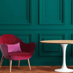 Poltrona rossa vellutata con cuscino rosa, accanto a un tavolino rotondo, su sfondo verde petrolio con pannelli decorativi.