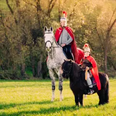 Zwei Reiter in römischer Kleidung, ein Mann auf einem Schimmel und ein Kind auf einem Pony, reiten im Sonnenschein. Ein Bild wie aus einer St. Martin-Geschichte.
