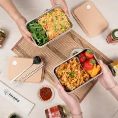 Two hands hold separate noodle bento boxes, one with fried rice and greens, the other with noodles, strawberries, and orange slices.