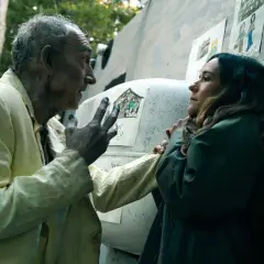 Un vieil homme aux cheveux gris, vêtu d'une chemise jaune, parle à une femme. Des dessins d'enfants ornent le mur derrière eux. Saison 5 date de sortie.