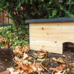 Ein neues Igelnest aus Holz steht im Garten, umgeben von Laub und Grün.