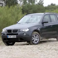 Ciemnoszary BMW X3 E83 pokonuje szutrową drogę, unosząc kurz.