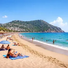 Słoneczny dzień na Kleopatra Beach. Ludzie opalają się na piasku i pływają w turkusowym morzu. W tle góra z zabudowaniami.