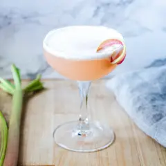 En rosa rabarber sour-drink med skum och en rabarberdekoration. Två stjälkar rabarber ligger bredvid.