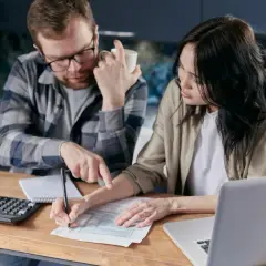Para wypełnia fakturę VAT, mężczyzna wskazuje na dokument, kobieta pisze. Obok kalkulator i laptop.