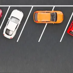 Cztery samochodziki: czerwony, biały, pomarańczowy i ponownie czerwony, zaparkowane na szarym tle z białymi liniami wyznaczającymi miejsca parkingowe.