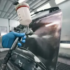 Ręka w niebieskiej rękawiczce trzyma pistolet lakierniczy, nanosząc podkład na element karoserii. Czas schnięcia podkładu epoksydowego zależy od warunków.