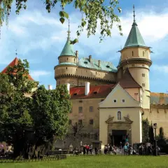 Bojnice zamek, z zielonymi wieżami i czerwonymi dachami, otoczony drzewami, z ludźmi spacerującymi po dziedzińcu.