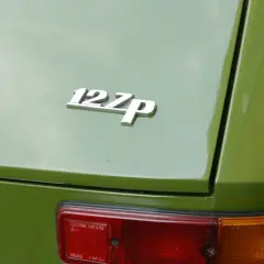 Chromowana plakietka "127p" na zielonej karoserii klasycznego Fiata 127. Widoczny fragment tylnej lampy.