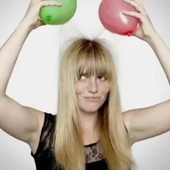 En kvinna med blont hår och fräknar håller upp en grön och en röd ballong ovanför huvudet, vilket får hennes hår att resa sig av statiskt elektricitet.