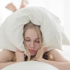 Kobieta z poduszką na głowie leży na łóżku, zamykając oczy.