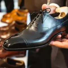 Main d'un homme tenant une chaussure noire brillante, prête à nourrir le cuir. D'autres chaussures en cuir sont alignées en arrière-plan.