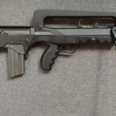 Czarny karabin FAMAS z charakterystycznym uchwytem i dwójnogiem, leżący na szarej tkaninie.