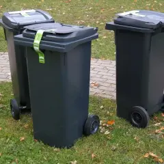 Trzy ciemnoszare kosze na śmieci stoją na trawie obok chodnika. Zastanawiasz się, gdzie wyrzucić piankę poliuretanową? Sprawdź lokalne przepisy dotyczące odpadów.