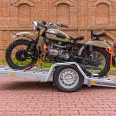 Motocykl Ural na przyczepie z pasami transportowymi, na tle ceglanego budynku.