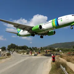 Ett flygplan landar lågt över vägen vid Skiathos flygplats. Människor står längs vägen och tittar upp.