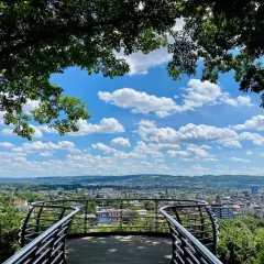 Blick vom Aussichtspunkt Wuppertal über die Stadt. Ein geschwungenes Geländer rahmt die weite Sicht auf Häuser und grüne Hügel unter einem blauen Himmel mit weißen Wolken ein.