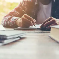 Osoba w kratę pisze w notatniku na drewnianym stole, obok książki i zeszyty.