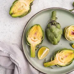 Un plato con una alcachofa entera y varias cortadas por la mitad, mostrando su interior amarillo y morado. También hay gajos de lima.
