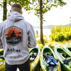Gotowy na spływy kajakowe? Na zdjęciu mężczyzna w bluzie z logo "Łap za wiosło" stoi obok rzędu kajaków nad jeziorem.