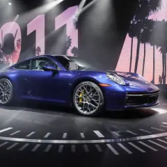 Un Porsche 911 992 azul metálico, con pinzas de freno amarillas, se exhibe sobre un escenario circular con un fondo de palmeras y el número 011.