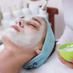 Une femme reçoit un masque ou gommage en premier, appliqué au pinceau sur son visage. Elle est détendue, les yeux fermés.