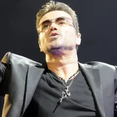 George Michael w okularach, w czarnej marynarce, śpiewa swoje ponadczasowe utwory.