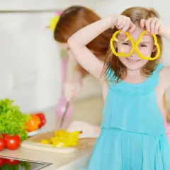 Dziewczynka w niebieskiej sukience bawi się plasterkami papryki, udając okulary. W tle siostra kroi warzywa. Zdrowa dieta, nawet przy celiakii, może być radosna!
