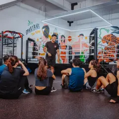 A boxing coach instructs a group sitting on the floor, preparing them for boxing reflex training. The gym features a boxing ring and punching bags.