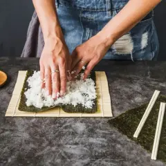 Ręce rozprowadzają ryż na nori, przygotowując zaprawę do sushi. Obok leżą pałeczki i marynowany imbir.