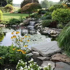 Ein idyllischer Garten mit einem kleinen Wasserfall, der in einen Teich mit Seerosen und bunten Fischen mündet. Ein perfektes Beispiel, wie man einen **Teich anlegen** kann.