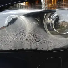 Zaparowane reflektory BMW z licznymi kropelkami wody wewnątrz. Widać odbicie domu i drzew.
