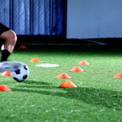 Piłkarz wykonuje trening reakcji, dryblując między pomarańczowymi pachołkami na sztucznej murawie.