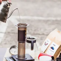 Parzenie kawy metodą Aeropress. Woda nalewana z czajnika do cylindra z kawą. Obok opakowania kawy i ziarna. Czas parzenia jest kluczowy.