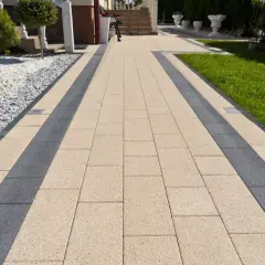 Eleganckie ścieżki z kostki brukowej w beżowo-grafitowym wzorze prowadzą do domu. Otoczone zielenią i białym żwirem.